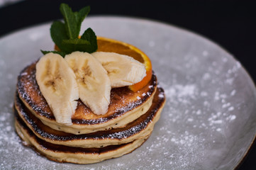 Homemade breakfast of pancakes decorated with orange, banana, mint and sugar powder on black background in grey plate.