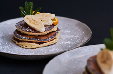 Homemade breakfast of pancakes decorated with orange, banana, mint and sugar powder on black background in grey plate.