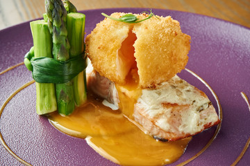An appetizing restaurant dish is chicken croquet with a flowing yolk on a salmon steak with a side dish of asparagus on a purple plate. Wood background. Selective focus.
