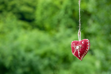 Isolated handmade cotton fabric heart hanging on bast string on green grass background. Red fabric...