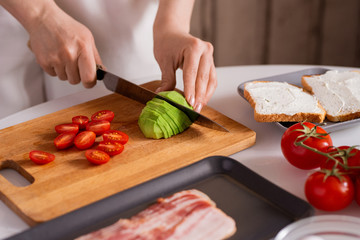Hands of housewife with knife cutting fresh avocado and tomatoes on board