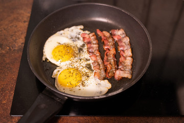 Frying pan with fried eggs and bacon on electric stove in process of cooking