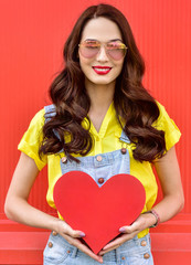 Beautiful woman holding a red heart over red background. Fashion portrait stylish pretty woman outdoor.