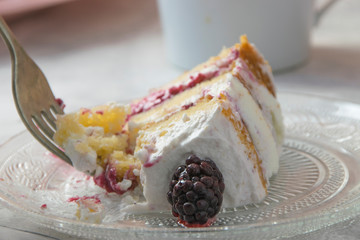 Fork taking a part of a slice of sponge cake filled with whipped cream and berries sauce with a blackberry as a decoration on a engraved crystal plate. Naked cake concept