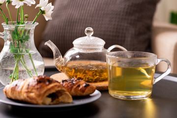 Teapot and cup with green herbal tea, fresh croissant on plate and flowers