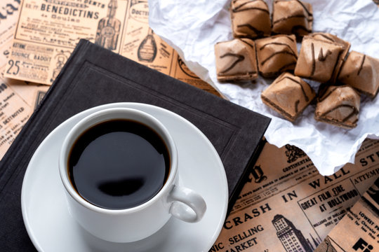 a cup of strong coffee on a white saucer and a black book on a table covered in old newspapers. cup with coffee and waffle cookies on white crumpled paper. vintage background from old newspapers
