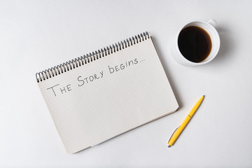 Notepad with words THE STORY BEGINS on white background. Top view of Notebook, Cup of coffee and pen