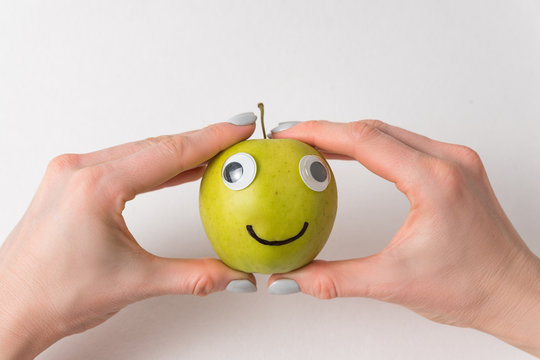 Hands holding cute little Apple with Googly eyes and drawn smile. Apple haracter on white background