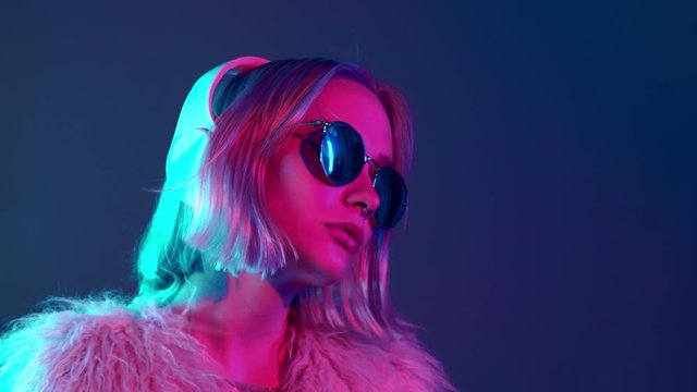 Glamorous hipster teenager in sunglasses and furry coat listening to music with headphones. Portrait of millennial pretty girl with short hairstyle with neon light. Dyed blue and pink hair.