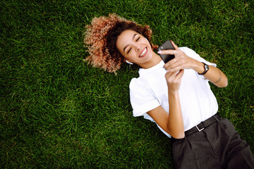 Young woman sitting on the grass  in a city park and using a smartphone. Technology and modern...