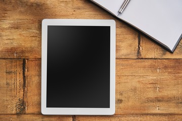 Blank screen white tablet on wooden table with open notebook. Flat lay.