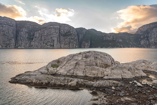 Stone island in the water of a fjord, surrounded by a mountain range at sunset