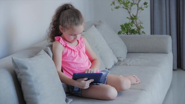 Curious cute kid little girl using digital tablet technology device included in studies sitting on sofa at home alone. Small child baby hold pad computer play games at home.Children tech addiction