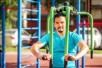 Fototapeta premium Young man in blue t-shirt doing chest exercises on butterfly training machine at public playground. Smiling man doing workout in sunny morning outdoors. Urban street background. Room for copy text