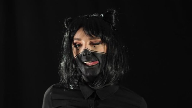 girl with black hair on a black background with black make-up, touches hair with fingers and shows tongue