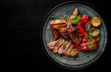 fried meat with potatoes, pepper, tomatoes, herbs and spices in plate on black wooden table background