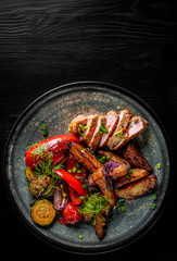 fried meat with potatoes, pepper, tomatoes, herbs and spices in plate on black wooden table background