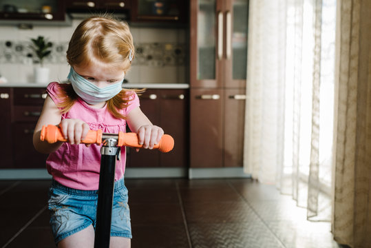 Kid wearing a mask. Little girl rides scooter. Happy child playing in kitchen during Covid-19 quarantine. Stay at home. Social media campaign for coronavirus prevention, hope during pandemic concept.