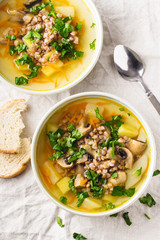 Soup of mushrooms, buckwheat, potatoes and carrots with herbs in bowls with two slice of white bread on the side, light background, vertical format.Top view