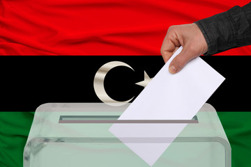 male voter drops a ballot in a transparent ballot box against the background of the Libya national...