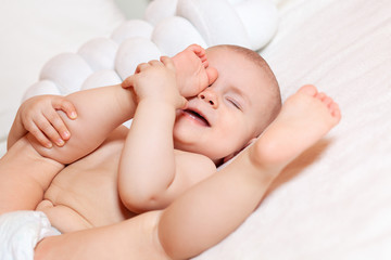 The baby is lying on his back on a white bed and playing funny. He raised his feet to his head and closed his eyes. Laughter, children's joy and fun. Products for children, toy.