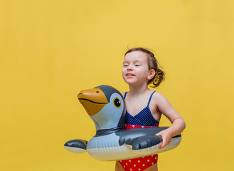 A little girl in a swimsuit with an inflatable penguin circle on a yellow background. Cute girl with pigtails in a polka-dot swimsuit. The girl smiles and looks away.