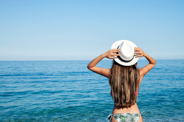 Fototapeta na wymiar Beach vacation. Hot beautiful woman in sun hat and bikini standing with her arms raised to her head enjoying looking view of beach ocean on hot summer day.