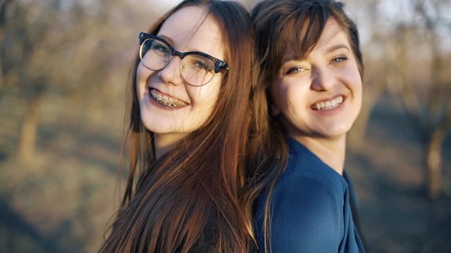 Two young girls,best friends or sisters, are joking, and laughing in a flowering garden.
One girl is standing with her back, another girl is looking forward.
They both have natural makeup.