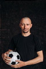 A male sports teacher stands against a dark background, holding a soccer ball