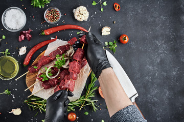 A butcher strands a fresh piece of meat for a barbecue on a bamboo skewer, four skewers of meat lie on a cutting board, which lies on a dark background. Top view, free space for text.