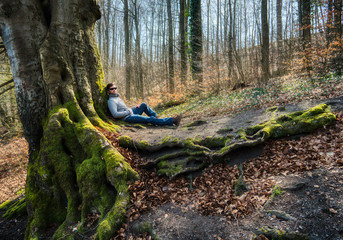young man in sunglasses relaxing at hte foot of a very old moss covered tree with gnarly roots in...