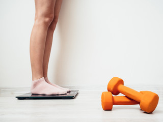 Female legs in socks stand on an electric scale against a white wall. Nearby are orange dumbbells for fitness. The concept of weight loss.