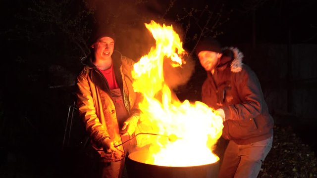 Two homeless young men are frying potatoes on a fire. Men stand at night near a barrel of fire and bask.