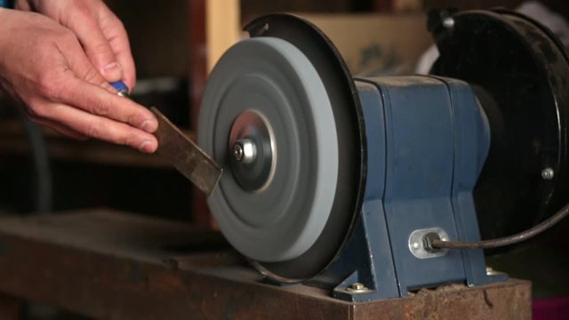 Faceless master sharpens chisel on grinding machine, sparks fly, male's hands holding woodworking tool, unknown person working indoor, electric bench grinder works, man sharpening chisels in workshop.