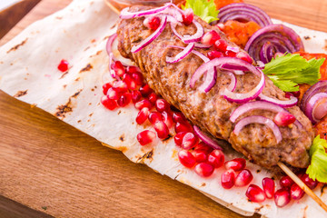 Kebab, meat on a skewer, pita bread, side dish of onions and tomatoes, pomegranate seeds and sauce on a wooden board