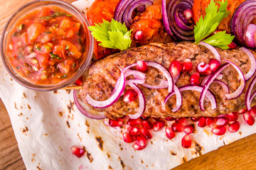 Kebab, meat on a skewer, pita bread, side dish of onions and tomatoes, pomegranate seeds and sauce on a wooden board