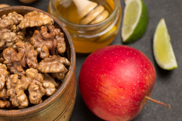 Walnut kernels in wooden bowl, honey in glass jar, lime slices and red apple on table.