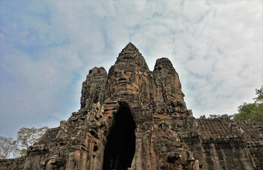 The gates of the ancient Angkor castle in Cambodia against a blue cloudy sky. On the ruins, human faces are carved from stone, looking to all sides of the world.