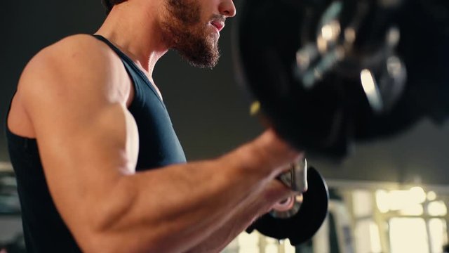 Close-up of face of handsome bearded young man with muscular wiry body wearing sportswear doing exercise with weights during sport workout training in modern dark gym. Tracking shot in slow motion.