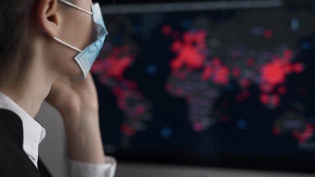 Side view close-up portrait of a young woman in disposable medical facial mask holding her head and looking at the epidemic map of the coronavirus Covid 19 pandemic on a computer