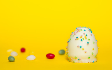 Fototapeta na wymiar Ein Osterei aus weißer Schokolade mit bunten Streuseln aus Zucker isoliert vor gelben Hintergrund, Symbol Ostern, Ei, naschen, Zucker