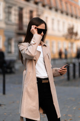 Beautiful brunette woman with phone walking on street.Fashion street style portrait. wearing dark casual trousers, white sweater,creamy coat and sunglasses .Fashion concept.Female business style.