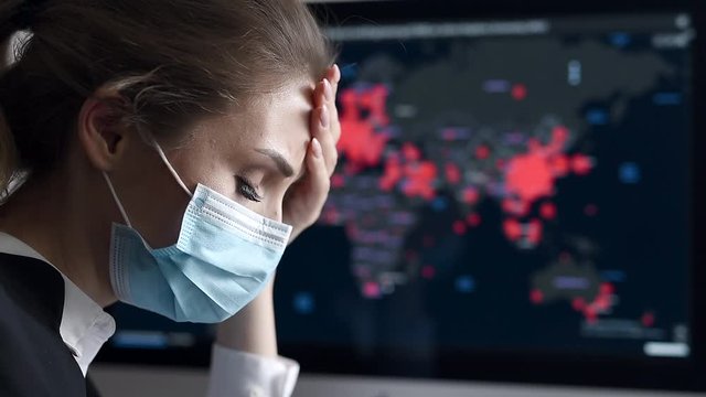 Side view close-up portrait of a young woman in disposable medical facial mask holding her head and looking at the epidemic map of the coronavirus Covid 19 pandemic on a computer