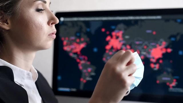 Side view close-up portrait of a young woman looks at the epidemic map of the coronavirus Covid 19 pandemic on a computer and puts on a disposable medical facial mask.