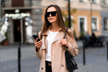 Beautiful brunette woman with phone walking on street.Fashion street style portrait. wearing dark casual trousers, white sweater,creamy coat. and sunglasses Fashion concept.Female business style.