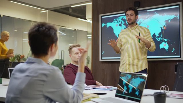 Tilt down medium shot of young manager presenting global market research standing by digital screen with world map, colleagues listening to him, arguing and asking questions in meeting