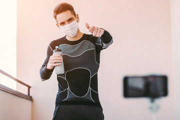 Confident man fitness instructor in medical face mask pointing to phone camera while exercising at...