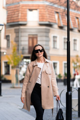 Fototapeta premium Outdoor photo of brunette lady walking on street background in autumn day.Fashion street style portrait. wearing dark casual trousers, white sweater,creamy coat and sunglasses.Fashion concept.