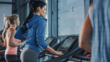 Fototapeta na wymiar Fit Athletic Woman Running on the Treadmill, Doing Her Fitness Exercise. Muscular Women and Men Actively Training in the Modern Gym. Sports People Workout in Fitness Center. Side View Shot