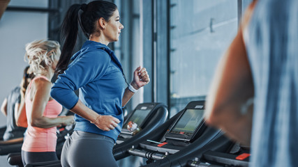 Fit Athletic Woman Running on the Treadmill, Doing Her Fitness Exercise. Muscular Women and Men Actively Training in the Modern Gym. Sports People Workout in Fitness Center. Side View Shot